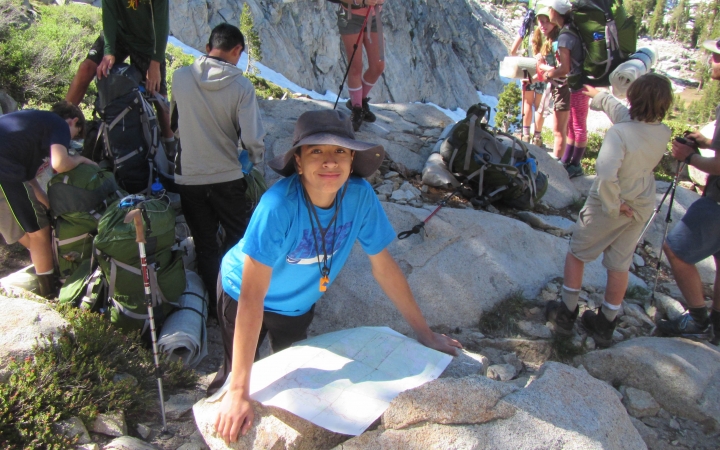 A young person leans against a rock on which a map is resting and smiles at the camera. Other outward bound students and their packs are pictured in the background. 